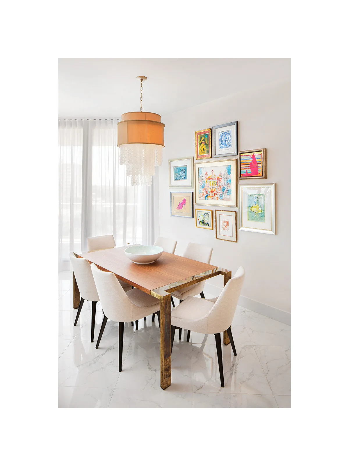 Small timber-topped dining table with pale chairs beside colorful framed pictures and an orange fringed pendant