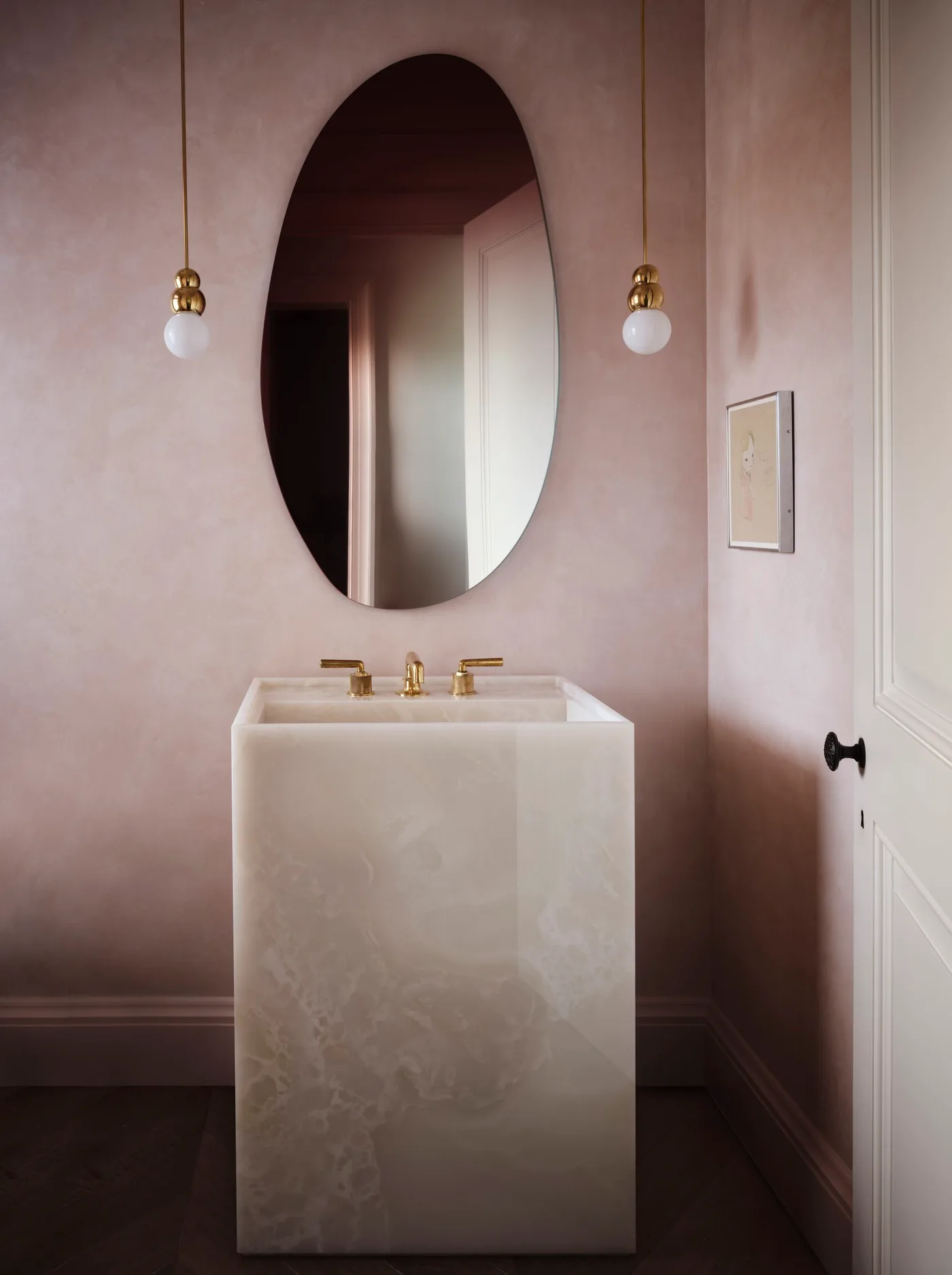 Pale rectangular basin pedestal against dusty pink walls beneath an oval mirror and two globe pendants