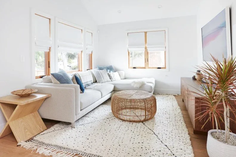 A white sectional and round woven table stand beside a low wood cabinet, pale rug and wood-trimmed windows with plain shades