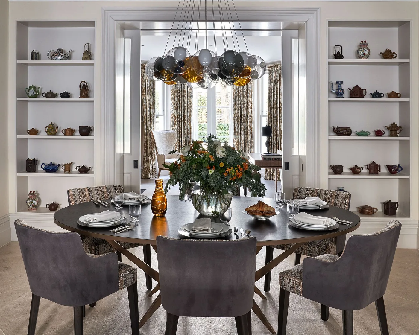 Gray upholstered chairs surround a round dark dining table beneath clustered glass shades beside pale shelves displaying small vessels