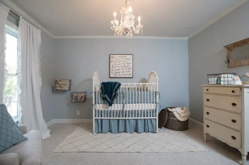 Blue-gray nursery with pale traditional-looking crib and dresser, white curtains, adult seating, baskets and decorative chandelier.