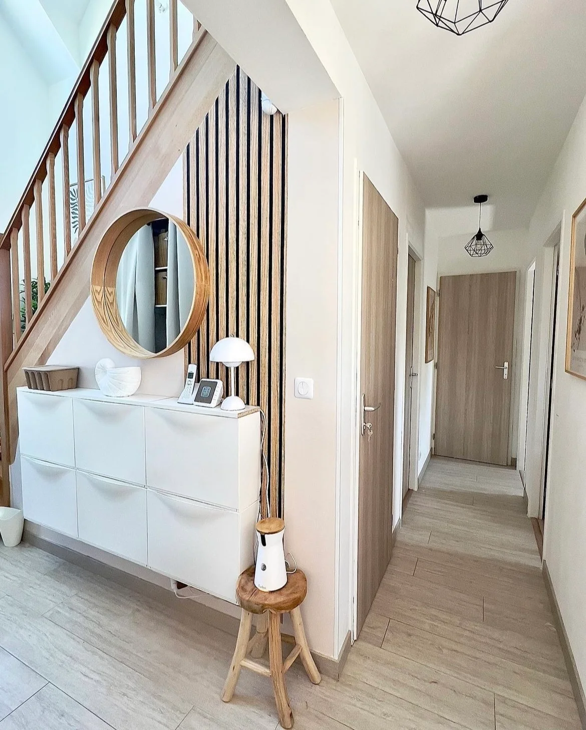 A white drawer cabinet stands against vertical wooden slats beneath a round wooden mirror beside a wooden staircase