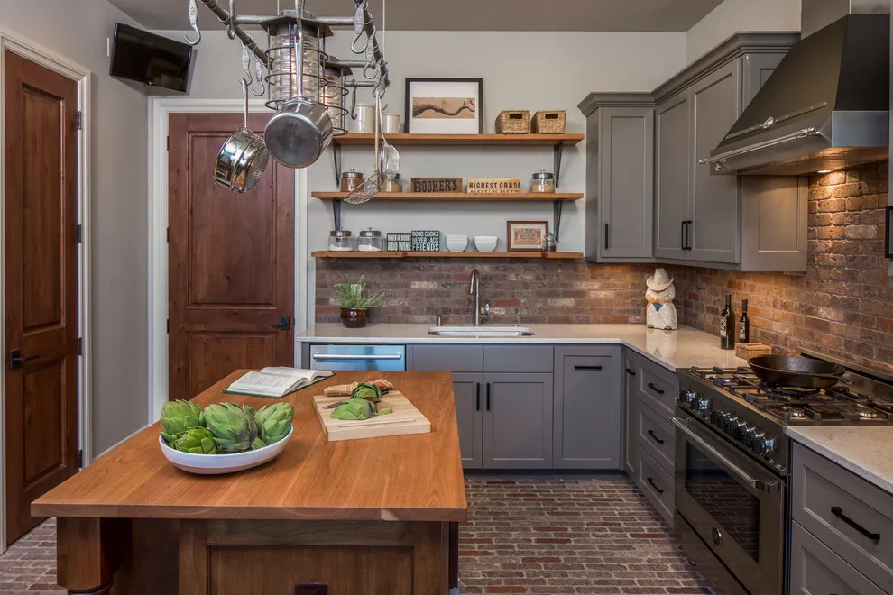 Gray painted cabinets sit beside a brick backsplash and a wooden worktable