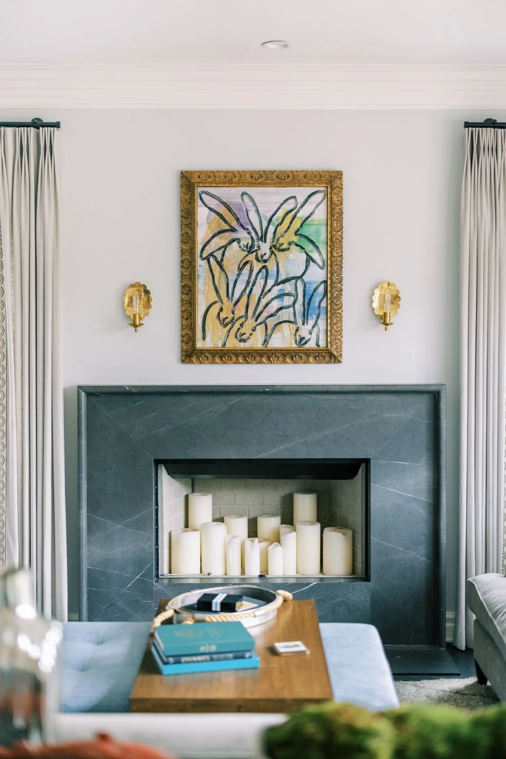 A dark green veined fireplace frame surrounds a pale recess with a group of unlit pillar candles beneath one colorful artwork and two small wall lights