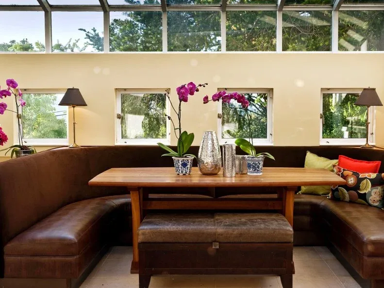 Dark brown wraparound banquette beneath high and low windows, with a long wooden table, an upholstered bench on the open side, orchids, and colorful pillows.