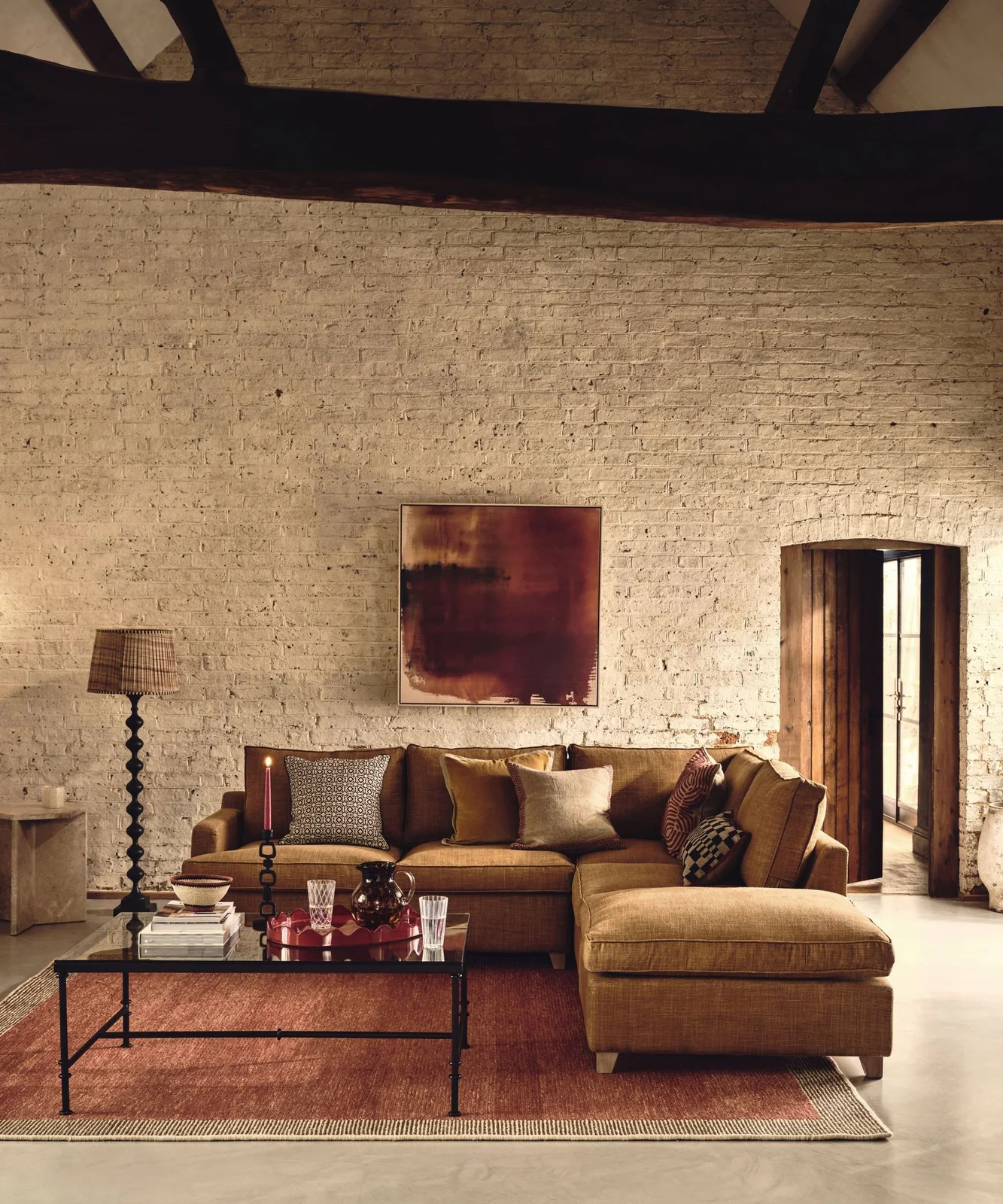 A brown sectional and red rug sit against pale brick living-room walls beneath dark beams beside patterned cushions and a slim dark coffee table