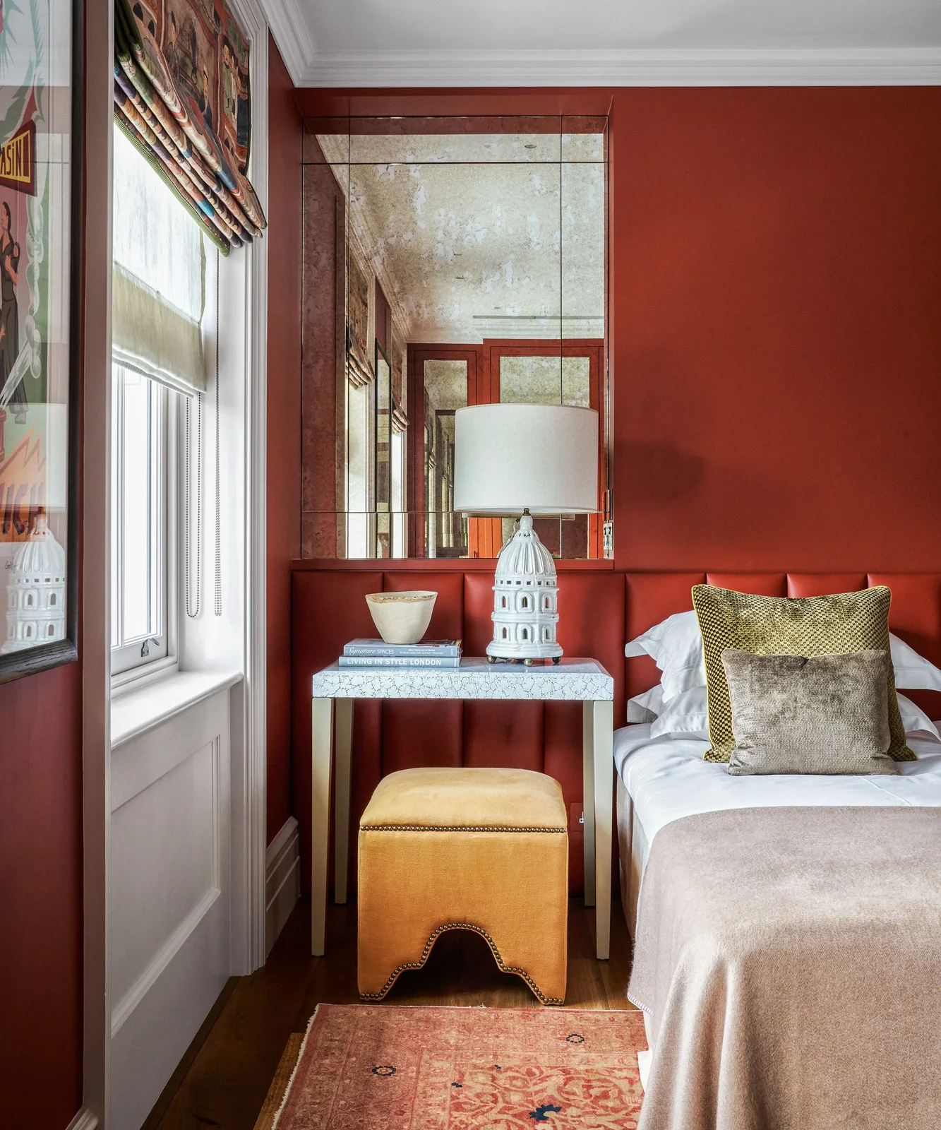 Deep red walls and a red channelled headboard surround pale bedding beside reflective wall panels, a floral blind and a yellow stool
