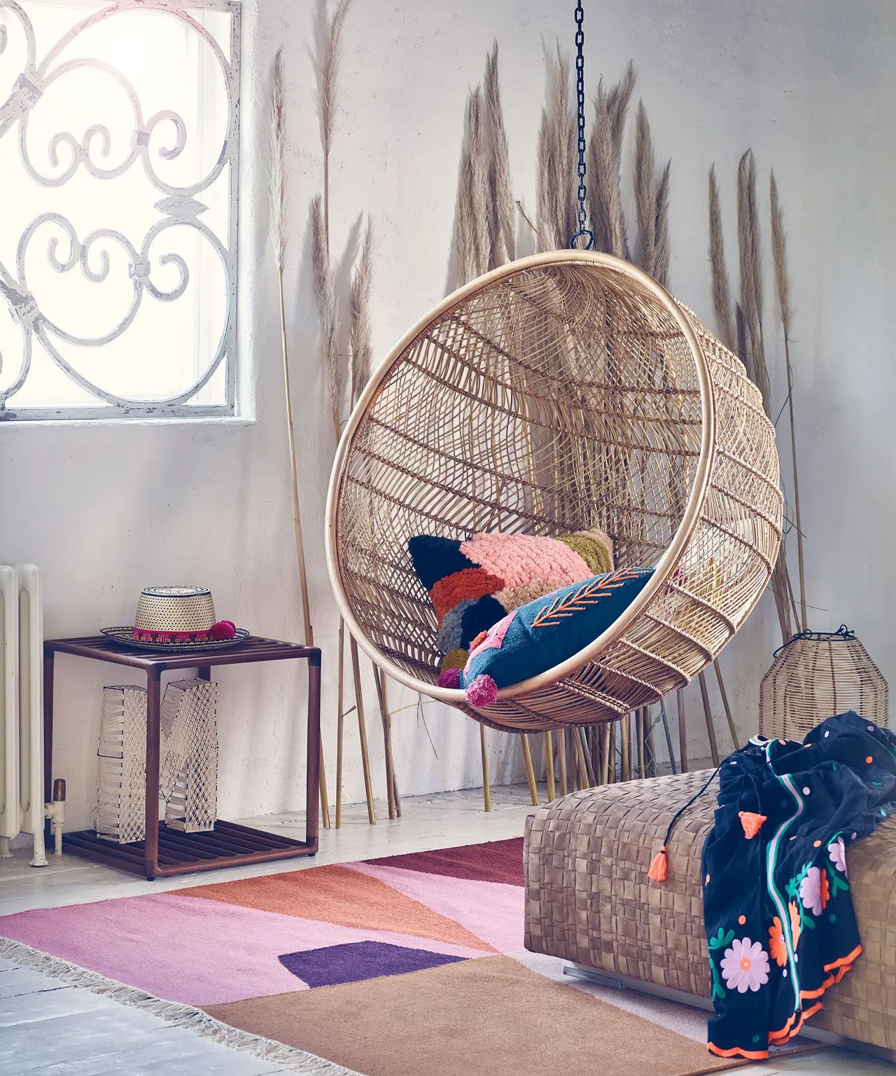 Bedroom corner with woven hanging chair, mixed colorful cushions, pink and purple geometric rug, woven floor basket and small dark side table.