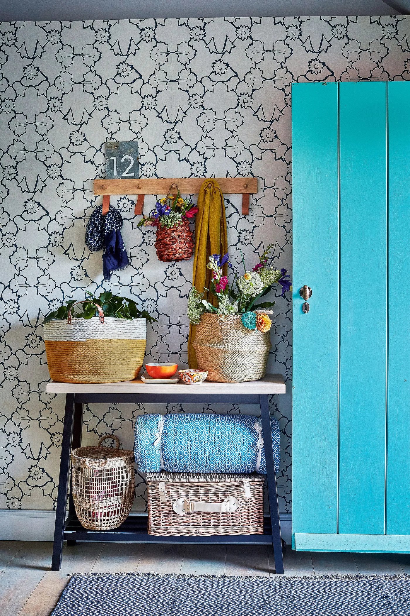 Country entrance detail with a turquoise door, pale dark-outlined floral wallpaper, wooden peg rail and a narrow dark console holding woven baskets.