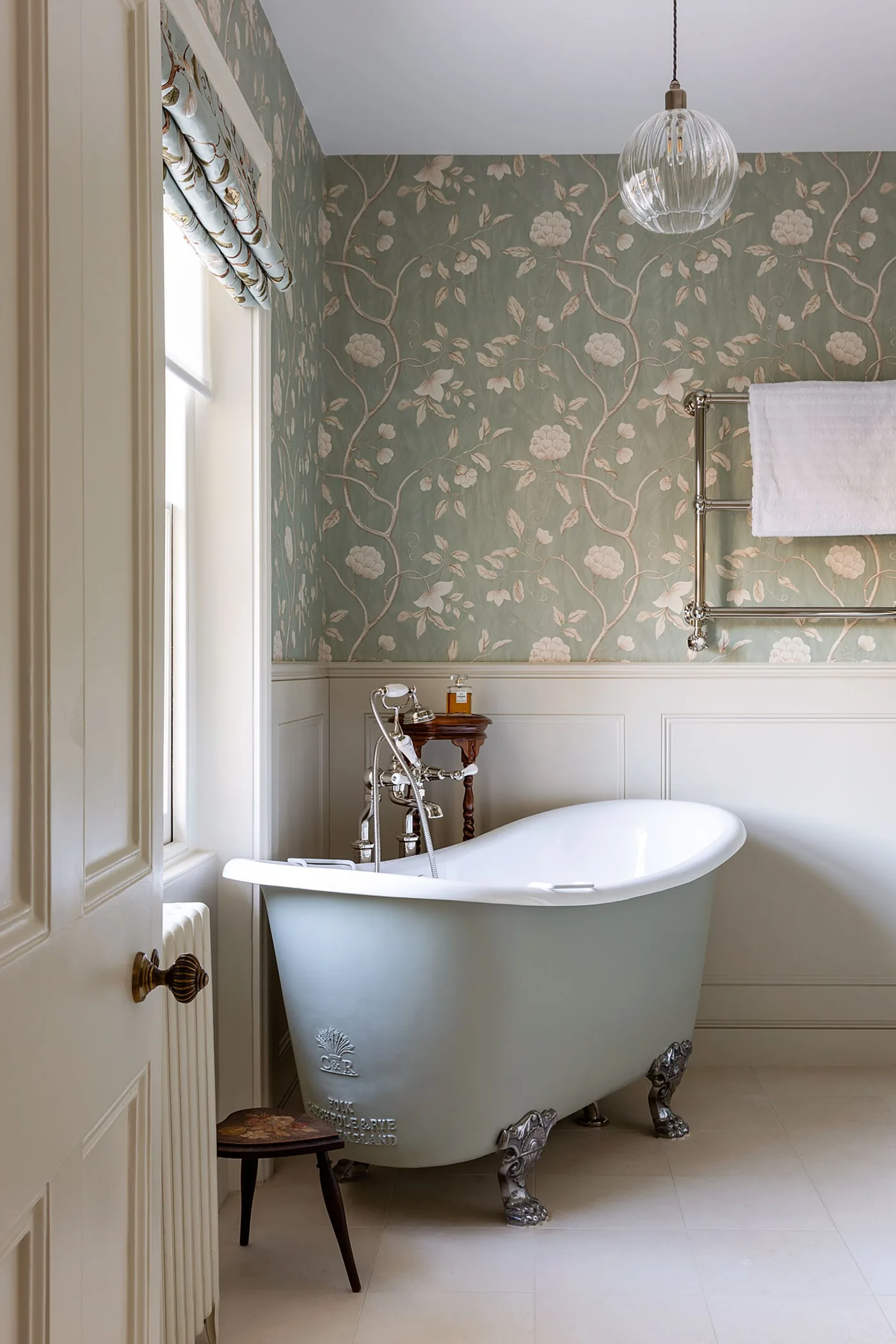 Country bathroom with a pale blue claw-foot bath, green floral wallpaper, pale lower paneling and a window beside the bathing area.