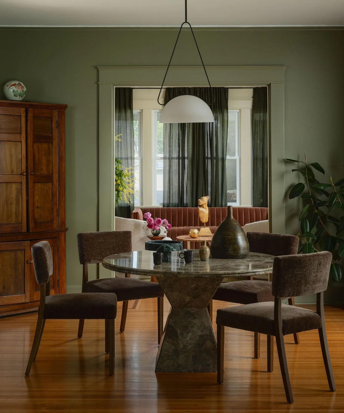 Dining room with green walls, a round dark veined table, brown upholstered chairs, white pendant shade and a wooden cupboard at the side.