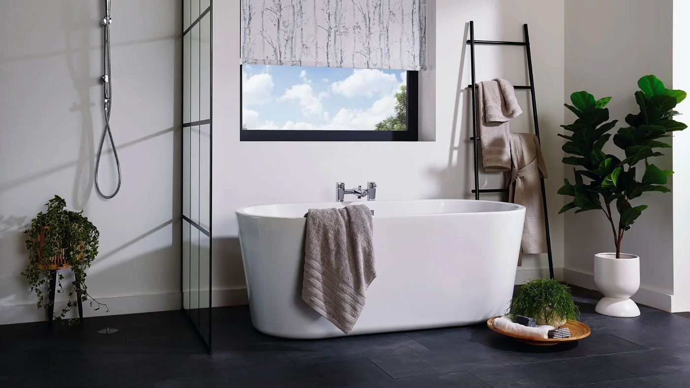 A white freestanding tub sits beneath a patterned window blind beside a black towel ladder, dark floor and leafy plant