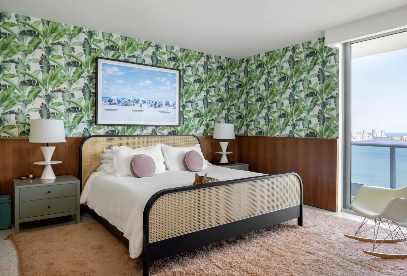 Green leaf wallpaper rises above wood bedroom paneling around a woven bed, white bedding, pale pink round cushions and matching bedside lamps