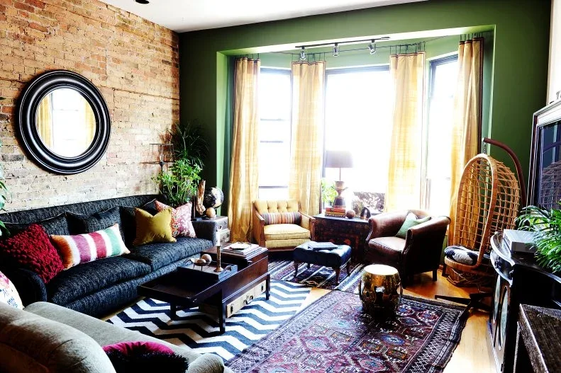 Living room with green wall around bay window, golden curtains, dark sofa, patterned cushions, woven hanging chair, exposed brick and mixed patterned rugs.