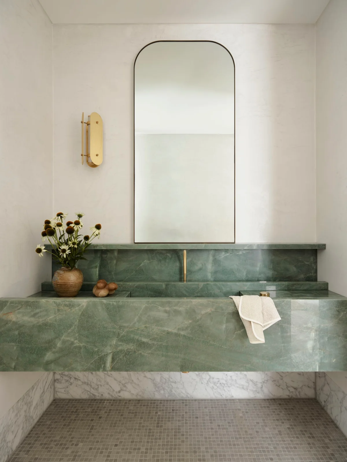 A broad green veined wall-mounted basin and backsplash stand beneath a tall rounded mirror against pale walls beside a slim warm-toned light