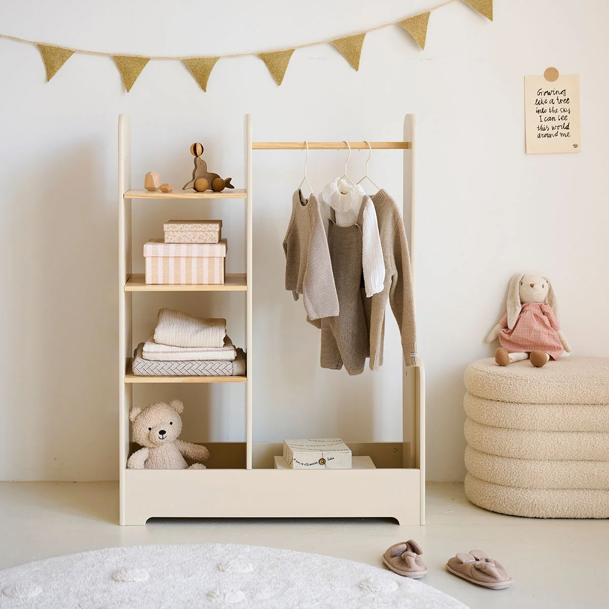 A white and wooden clothes rail with a lower drawer holds small garments beside shelves and a round gray pouf