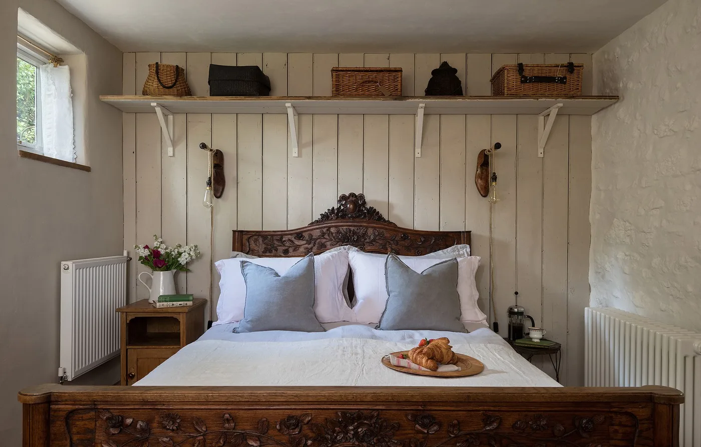 Country bedroom with an ornate wooden bed frame, pale blue and white bedding, vertical wall boards and a high shelf holding baskets and bags.