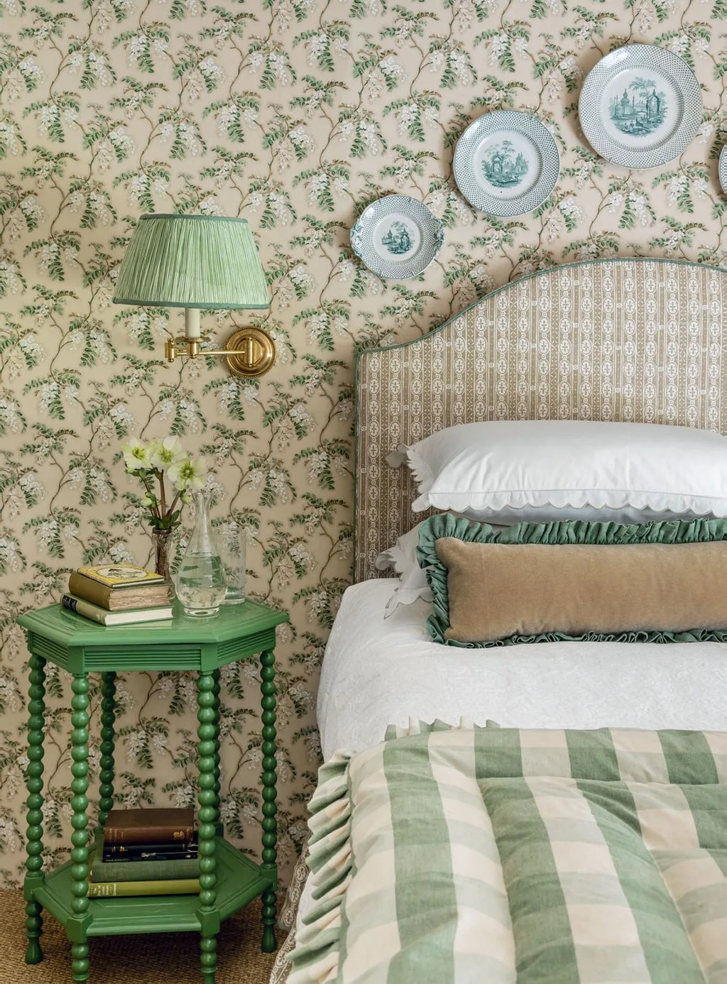 Bedroom with green leafy wallpaper, shaped pale headboard, green checked quilt, green wall shade, turned bedside table and decorative plates