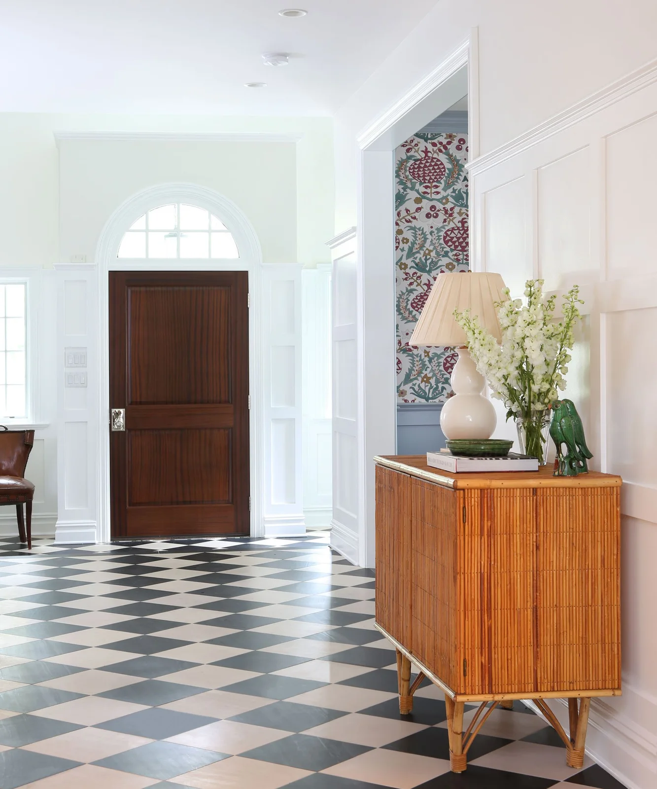 A hallway has dark-and-pale checked flooring beneath white paneled walls beside a warm wood cabinet and a dark wood front door