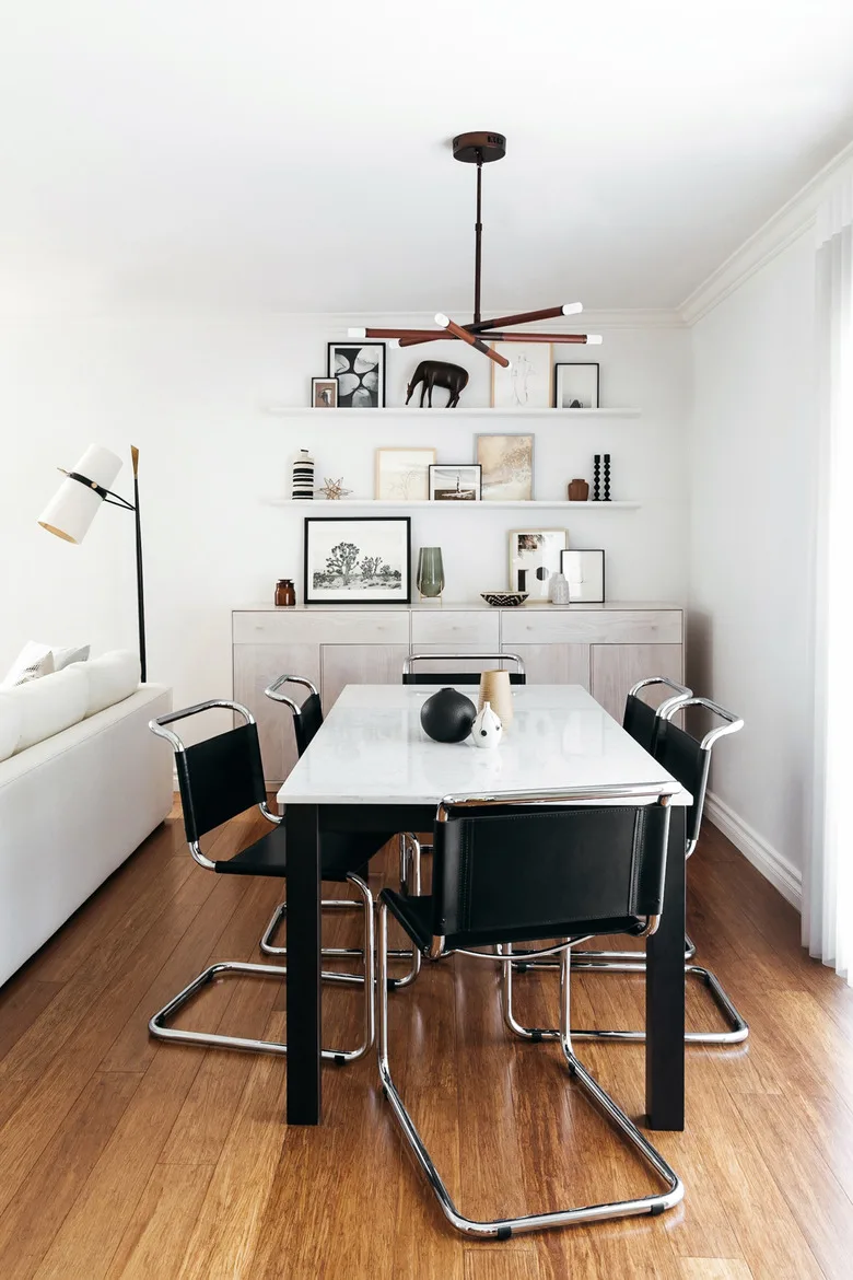 Two white floating shelves with framed pictures above a pale sideboard behind a white dining table and black chairs