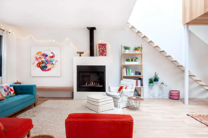 A plain white fireplace surround and black opening stand beneath a dark flue beside colorful chairs, a blue sofa, wood shelving and a light staircase