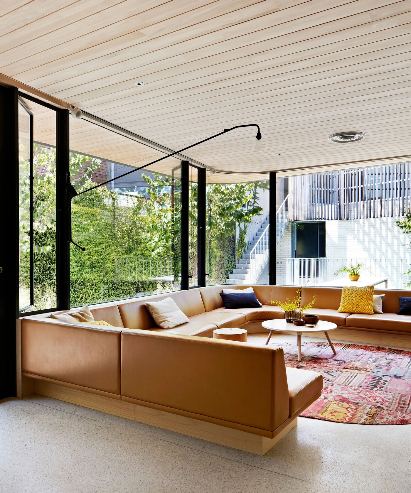 Living room with large low tan sectional, pale wooden ceiling, broad windows, patterned pink rug and light round central table.