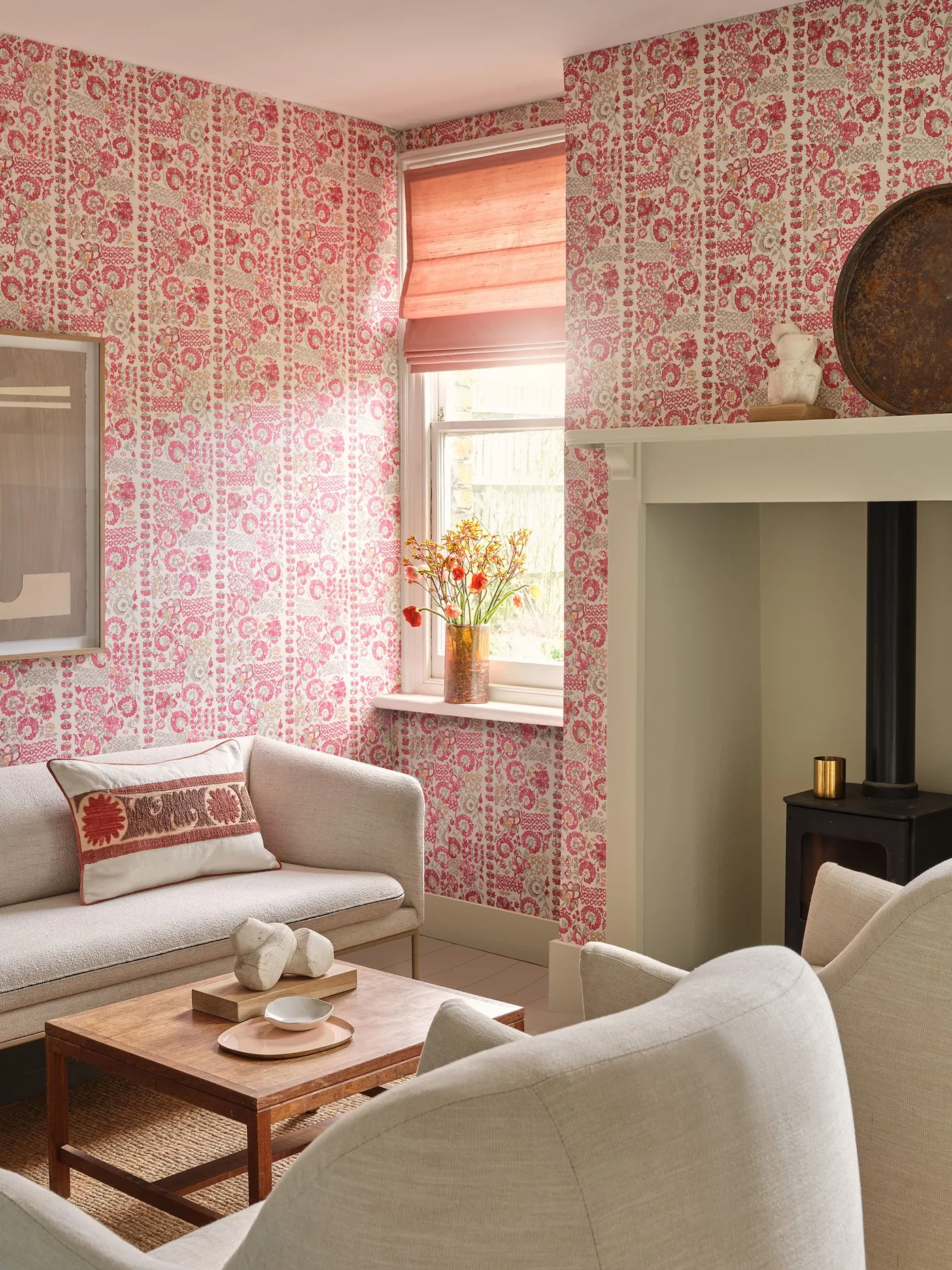 Sitting room with pink patterned walls, pale sofa and chair, wooden coffee table, pink window blind and simple fireplace surround.