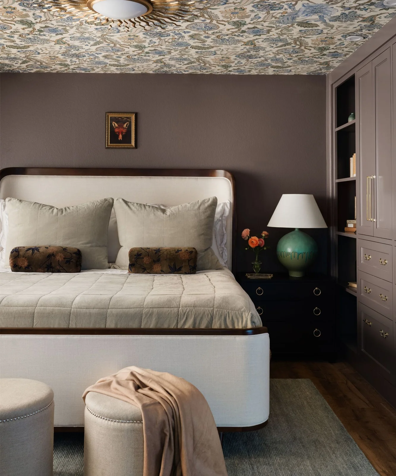 A dense botanical ceiling sits above brown walls, pale upholstered bed ends, patterned bolsters and two round pale stools