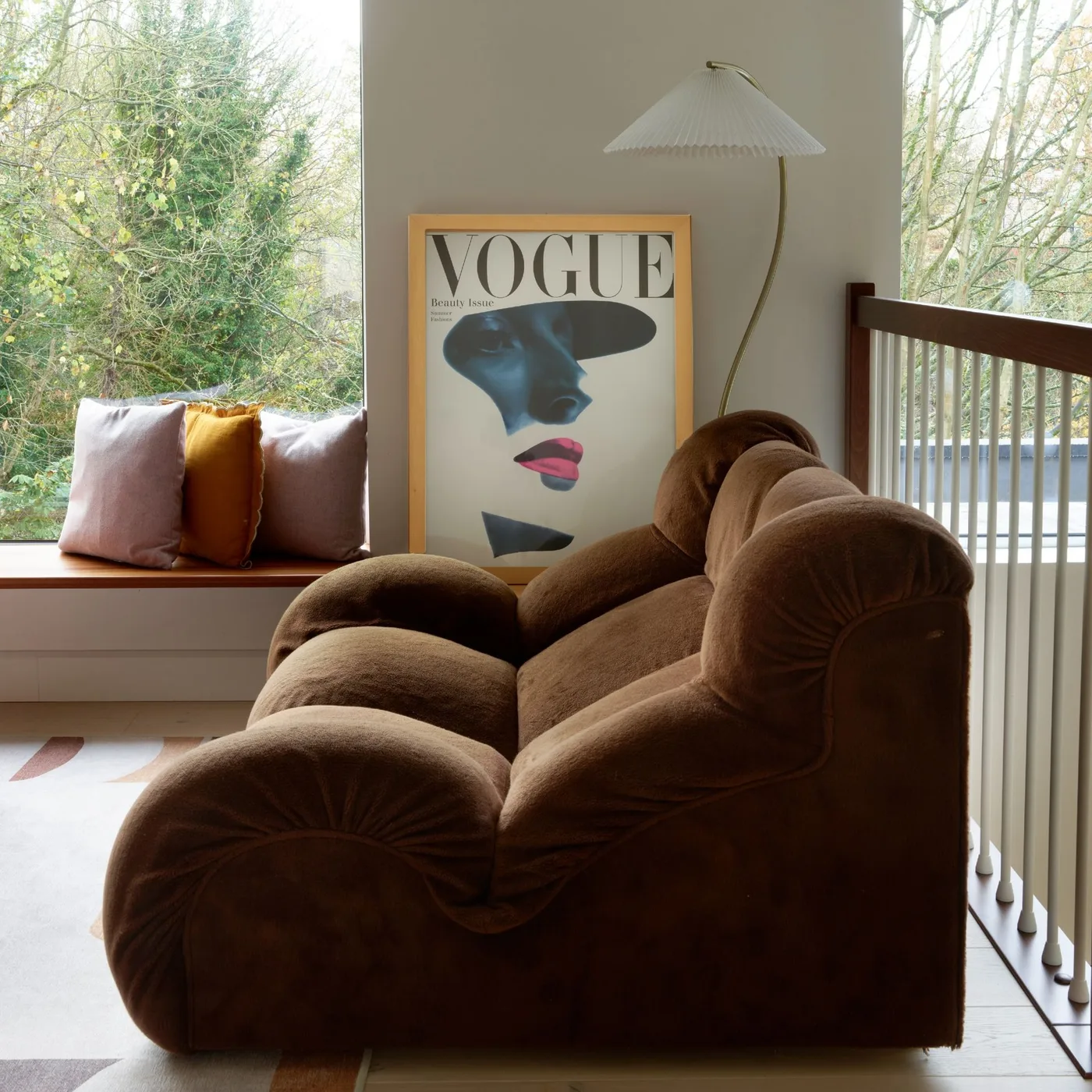 A sculptural brown sofa sits beside broad windows, a slim curved floor lamp, a window bench with cushions and one large framed print