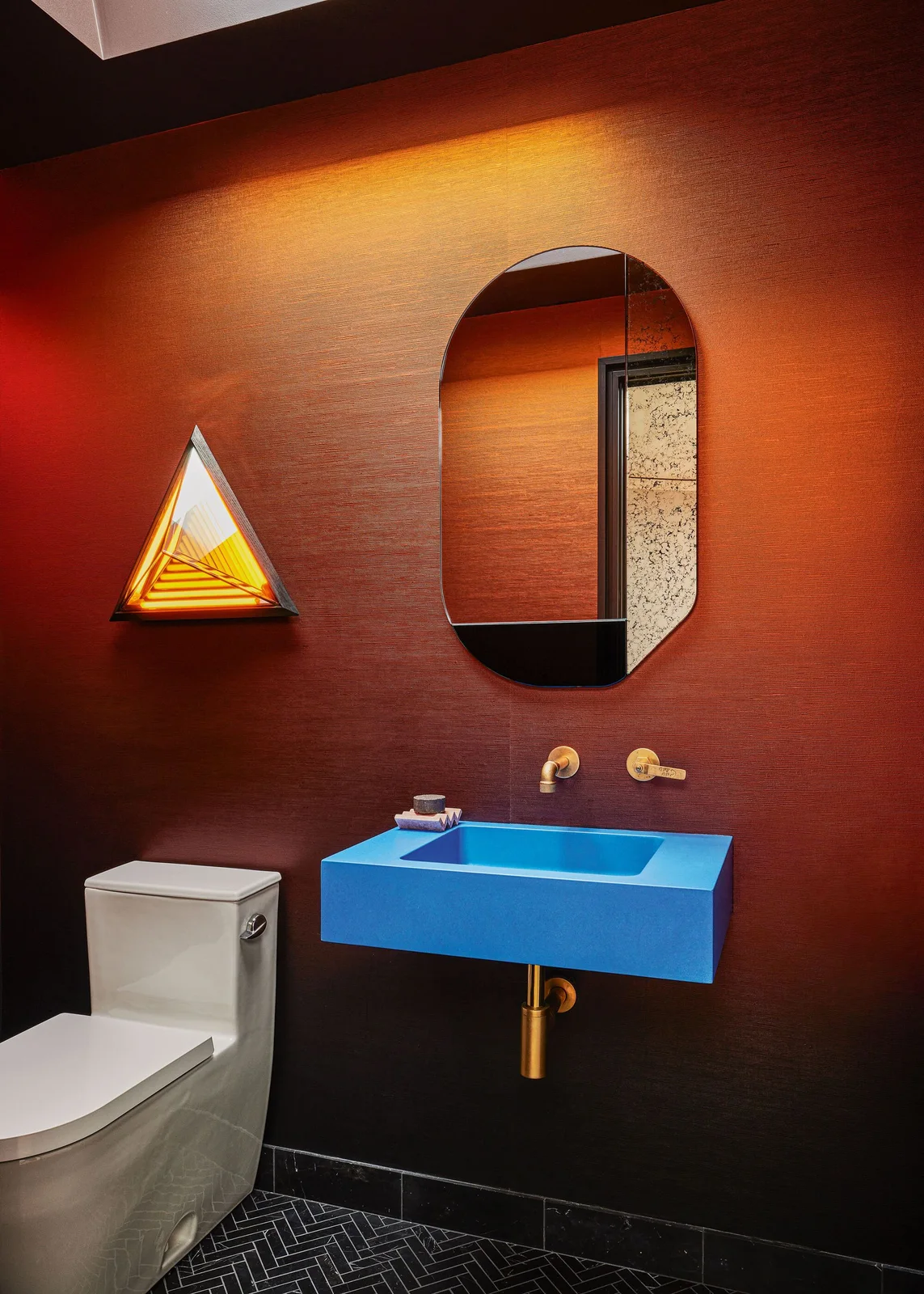 A bright blue rectangular wall basin stands beneath an oval mirror and warm-colored walls beside a triangular light, white toilet and dark patterned floor