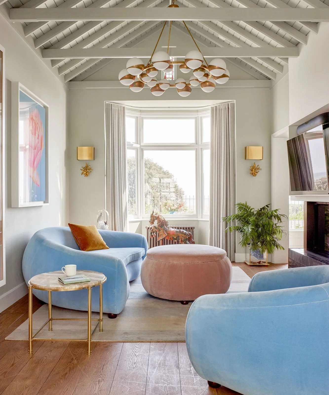 A television is angled beside curved pale blue chairs and a pink ottoman beneath a decorative light and white roof beams