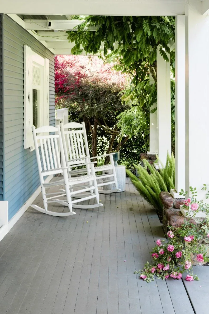 Two white rocking chairs on gray farmhouse porch beside flowering shrubs and ferns