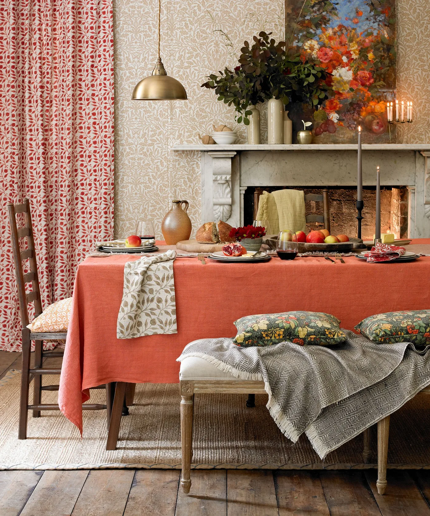 An orange tablecloth sits beside red-and-white leafy curtains, pale botanical wallpaper, floral cushions and a colorful painting above a fireplace