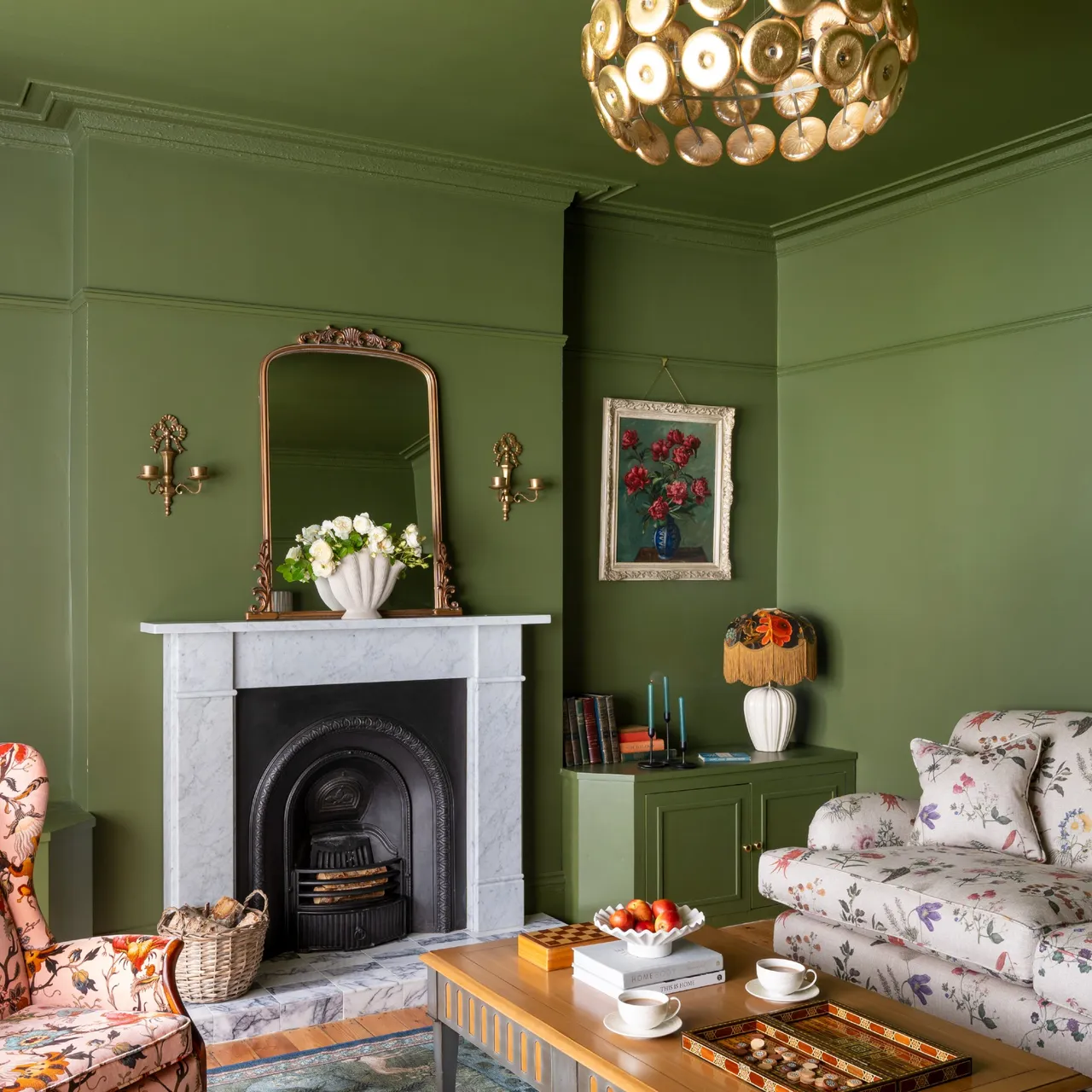 Green walls and cabinetry surround floral seating and a timber table beside a white fireplace