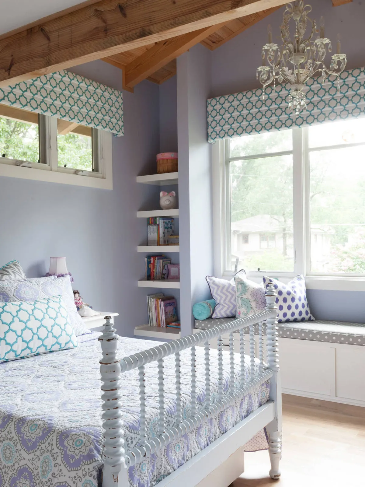 A pale spindle bed sits beneath timber ceiling beams beside a cushioned window seat.