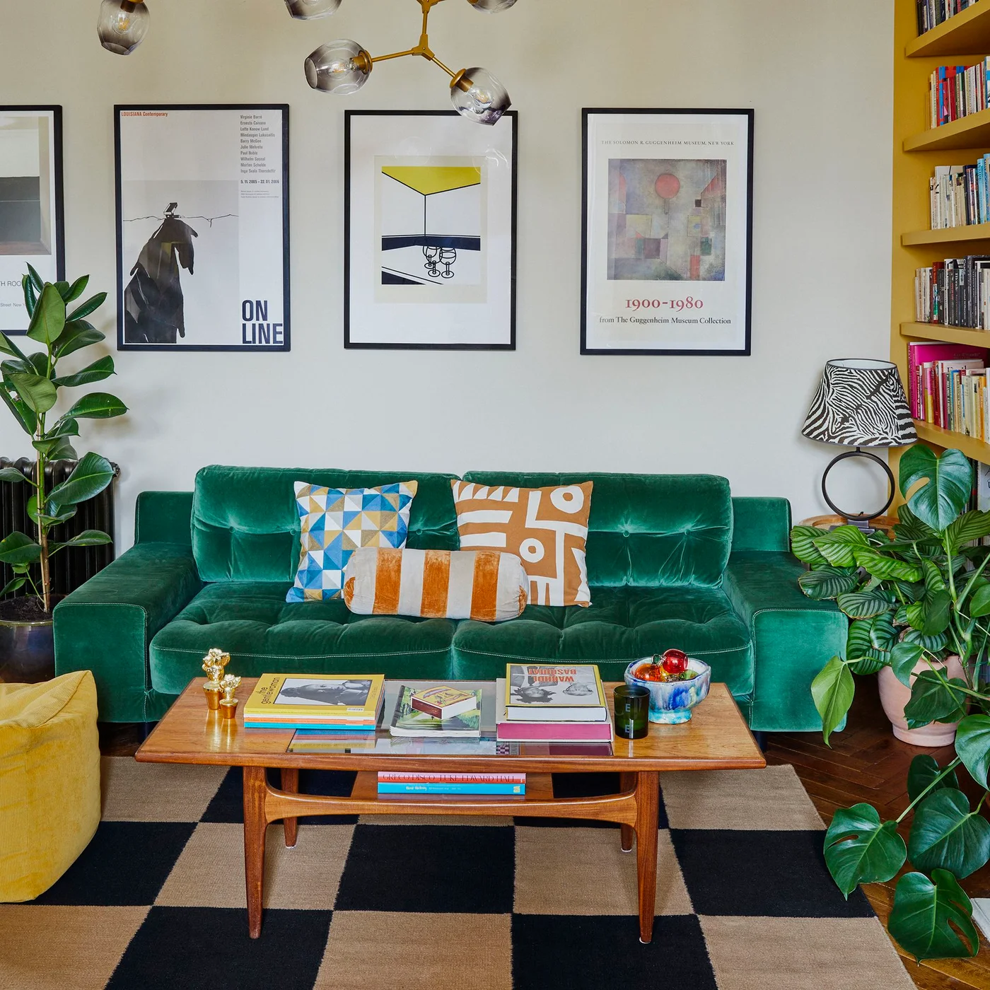 A buttoned green sofa has geometric cushions beneath framed posters beside a yellow bookcase and wooden table