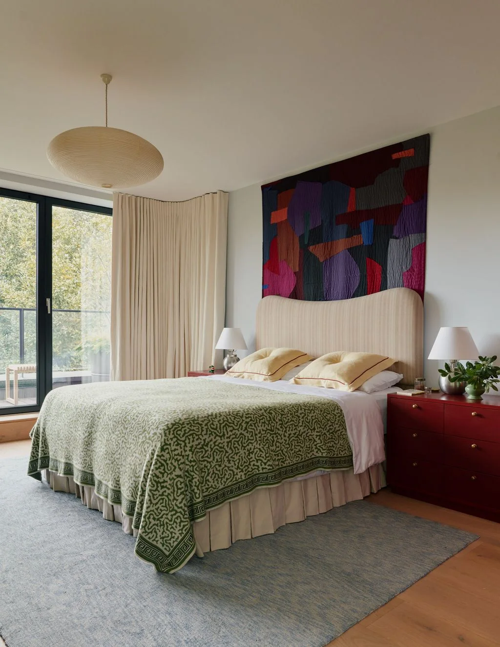 A pale upholstered bed with a green patterned cover stands below a colorful wall hanging beside red drawer chests, pale lamps and light curtains