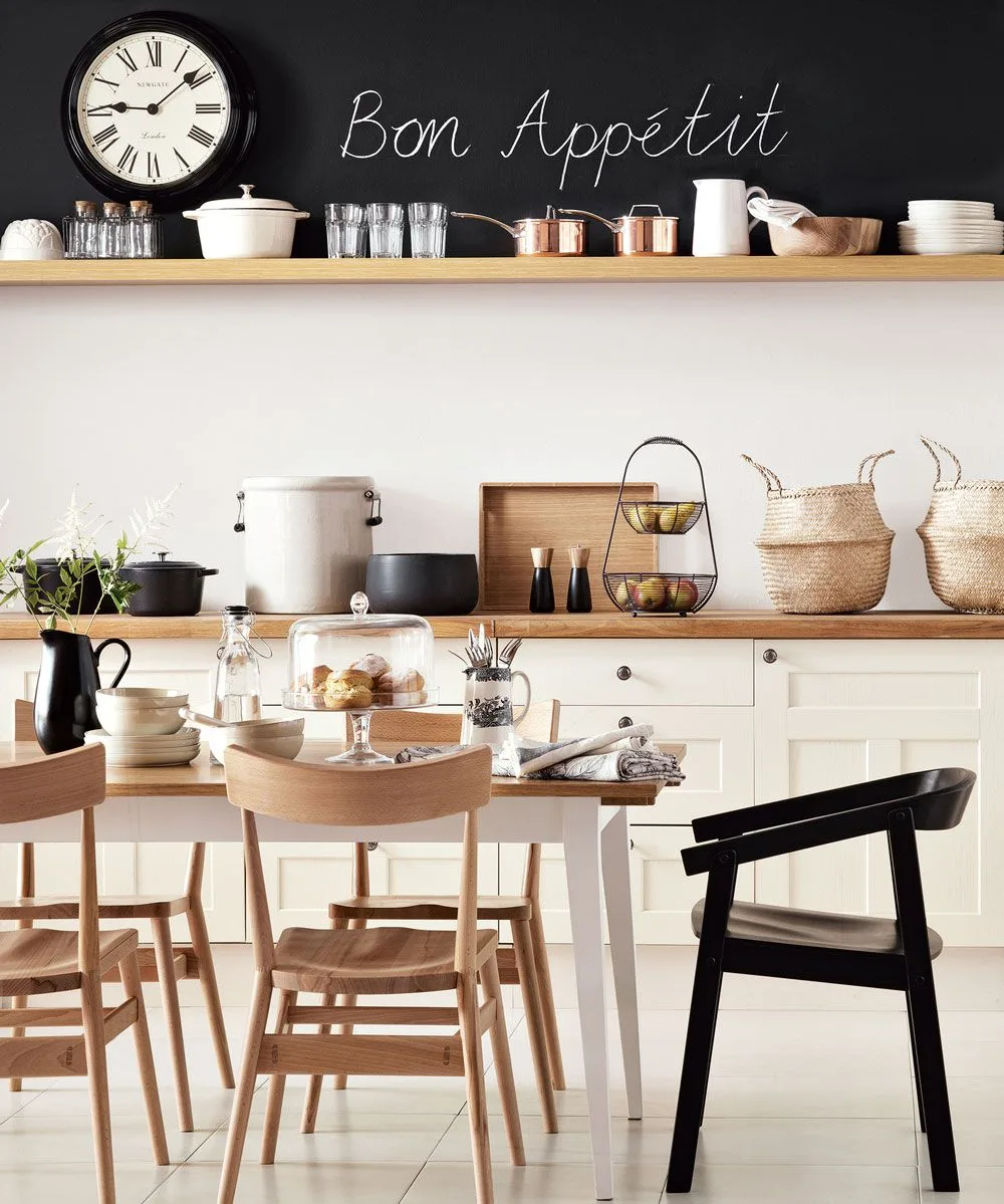 Cream lower cabinets sit beneath wooden shelves and a black chalkboard strip beside a dining table