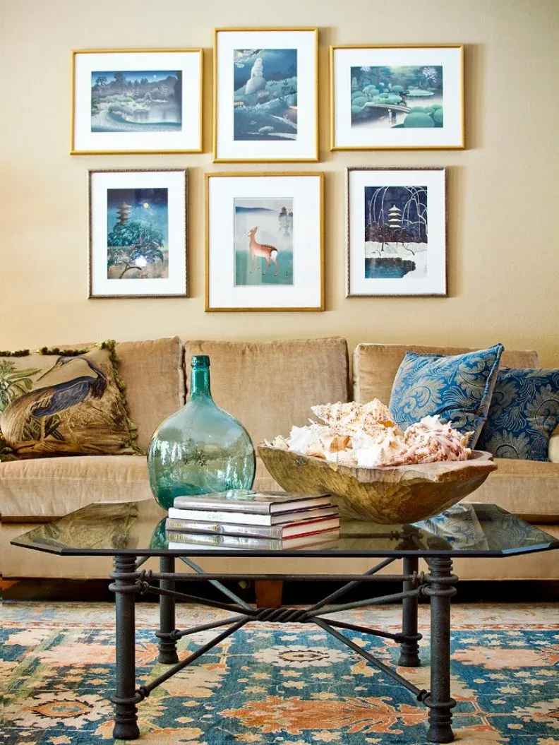 Tan sofa beneath six gold-colored frames with blue pictures, a blue cushion and a green glass vessel.