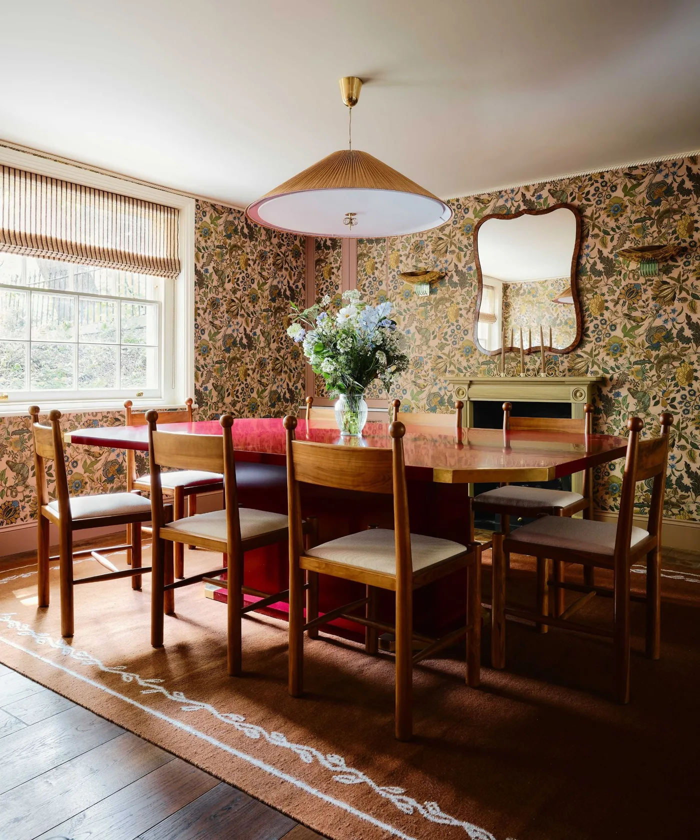 Cottage dining room with floral wallpaper, red table, wooden chairs with pale seats, a broad pendant shade and a curved wall mirror.