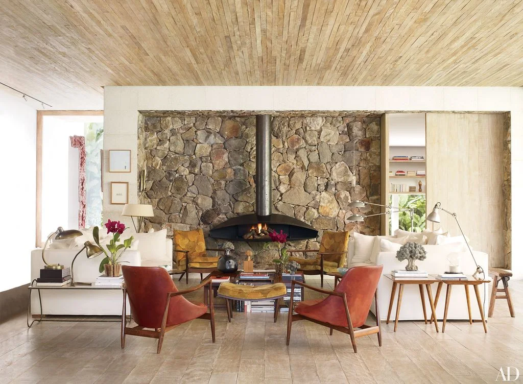 An angular black fireplace hood and vertical flue stand before a rough stone wall beside red and yellow chairs and pale sofas