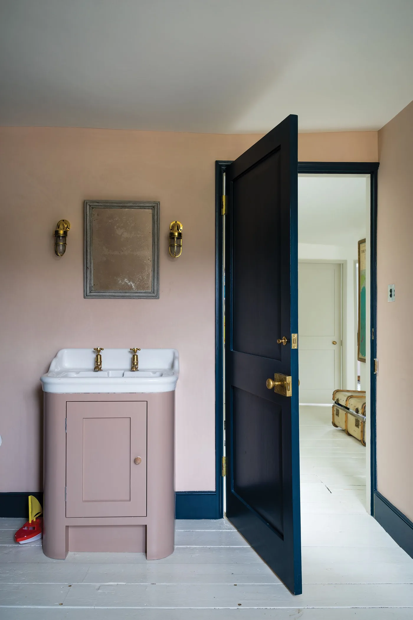 Small pink vanity and pink walls beside a deep blue door, with a framed mirror and two wall fittings.