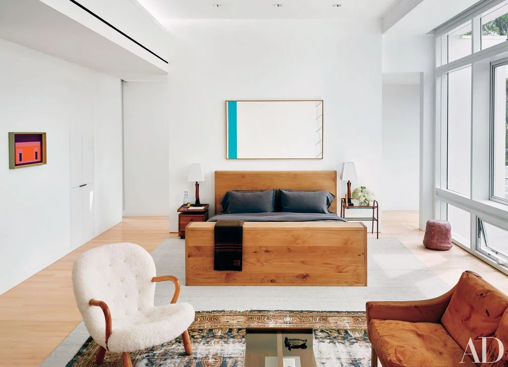 A plain wood bed with dark bedding sits beneath simple artwork within tall white walls beside broad windows, pale flooring and a small sitting group