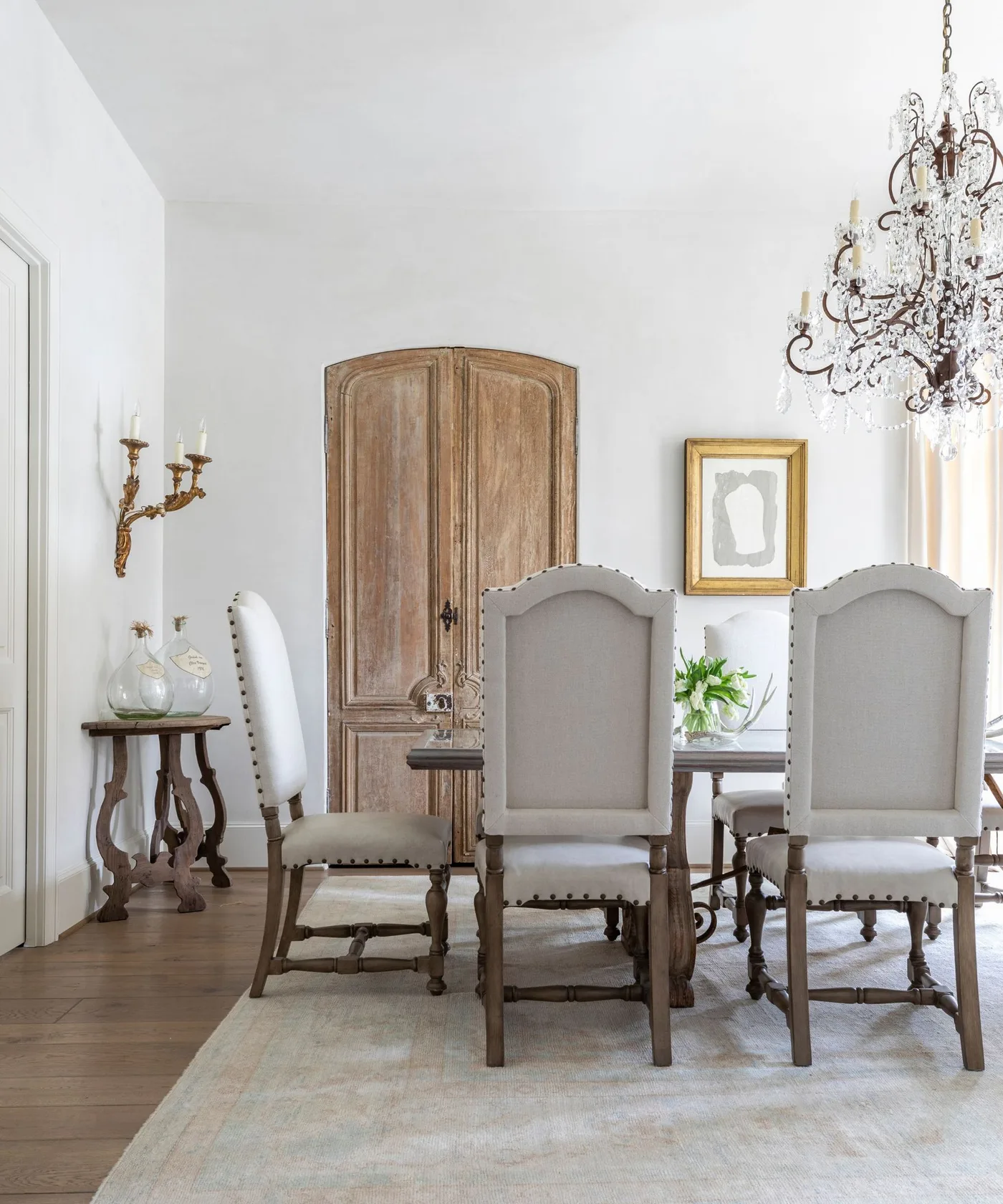 Pale high-backed dining chairs with decorative wooden frames beside arched wooden doors and an ornate chandelier.