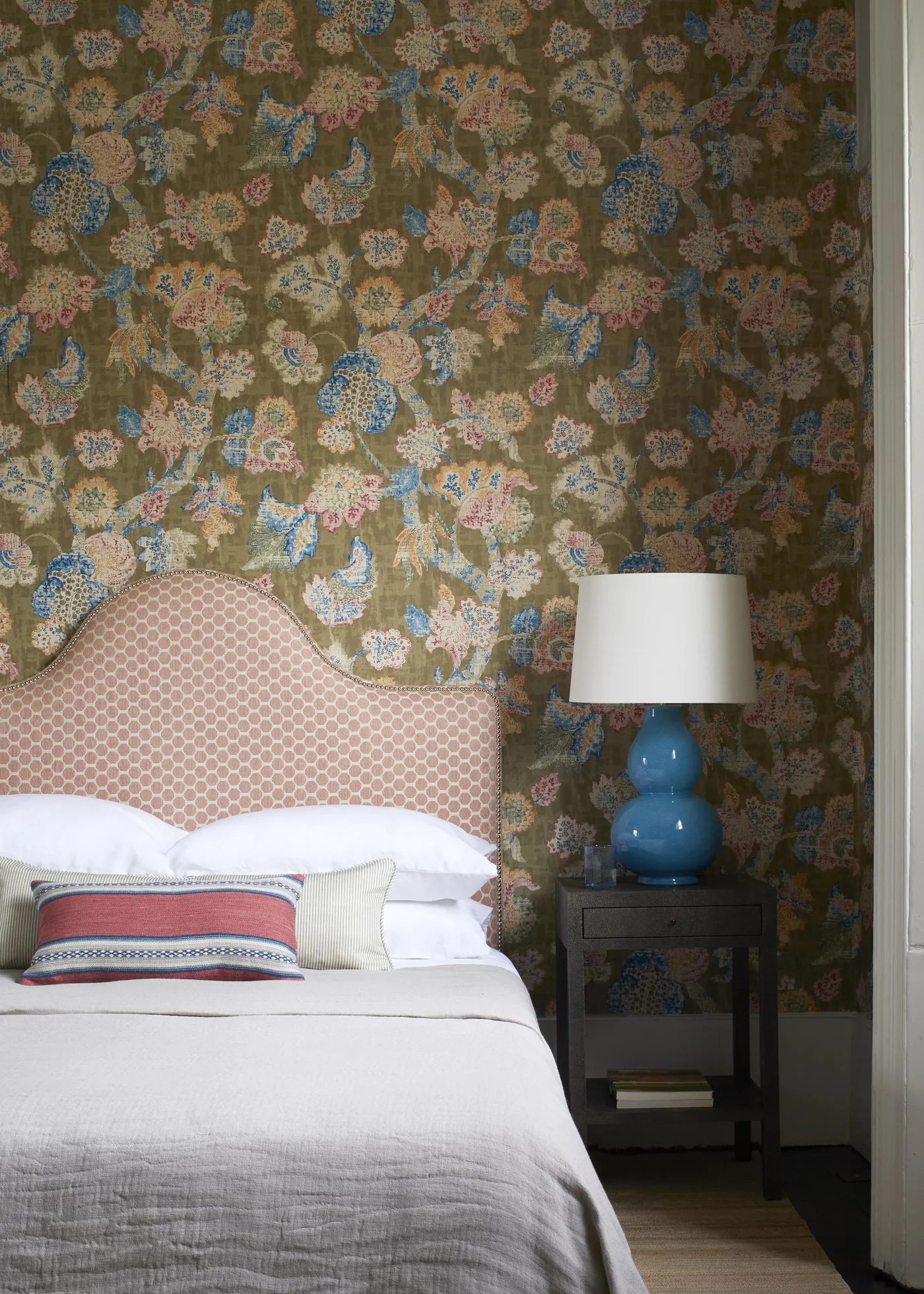 Dense muted floral wallpaper behind a pink curved headboard and a blue bedside lamp.