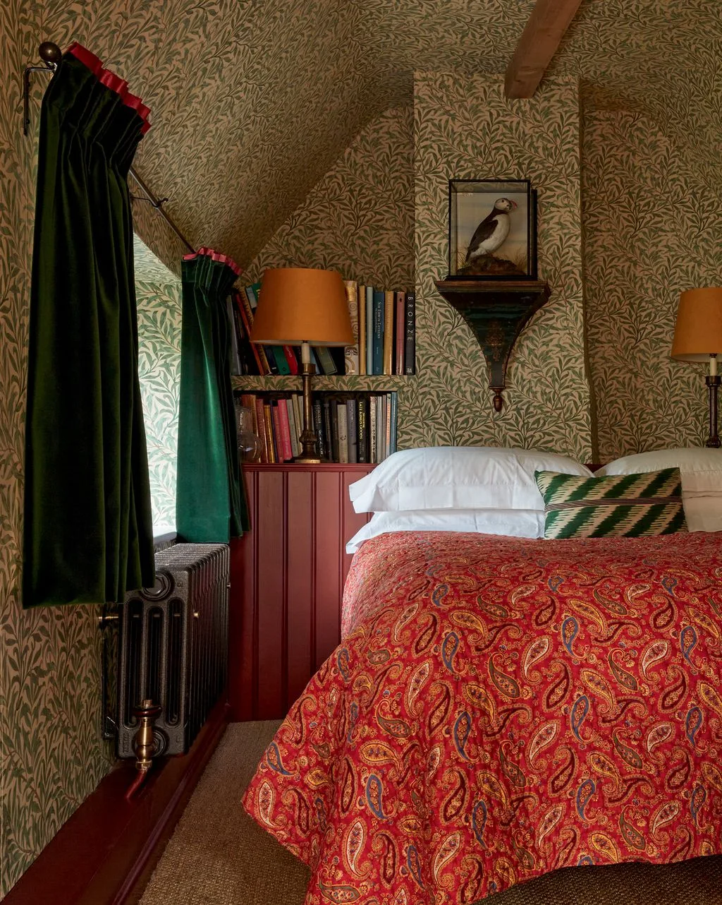Attic bedroom with leafy patterned walls and ceiling, dark green curtains, red paisley cover, small bookshelf and orange lampshade