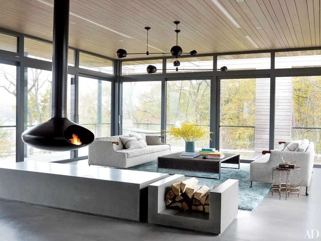 A black suspended fireplace hangs beside tall windows and pale sofas above a dark table and blue-green rug