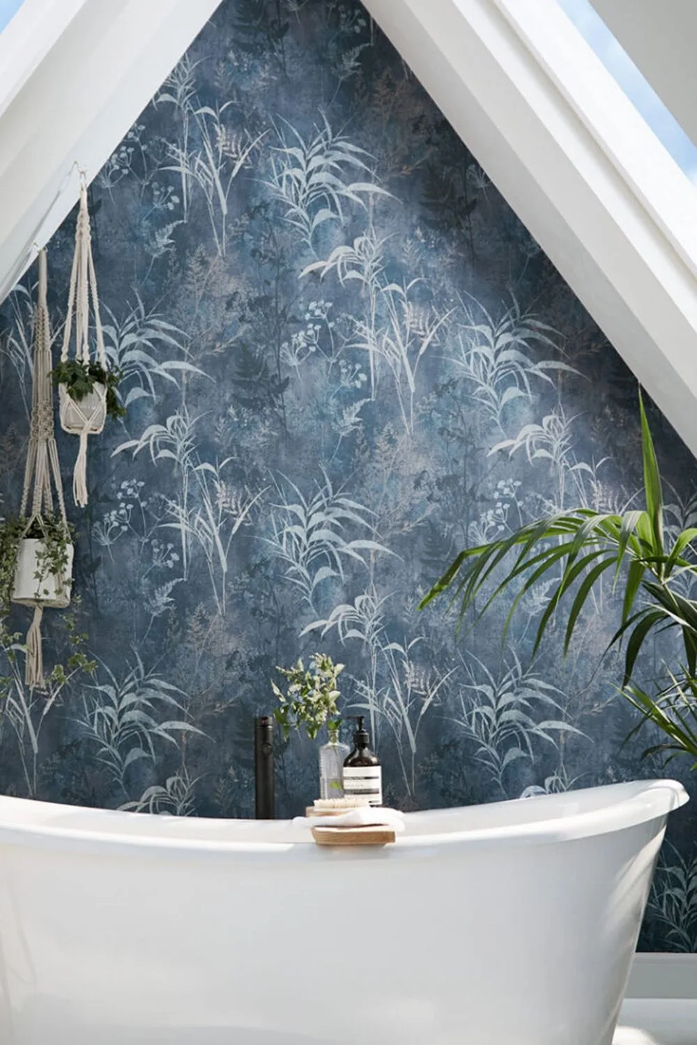A white freestanding bath sits against dark blue botanical wallpaper beneath a sloping ceiling