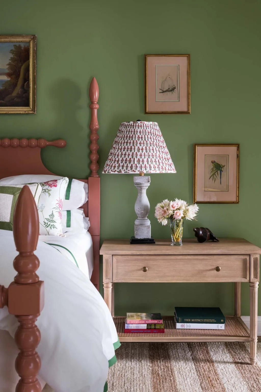 Olive green bedroom with a wooden spindle bed, pale floral bedding, open-shelf bedside table, bird artwork, and lamp with patterned shade.