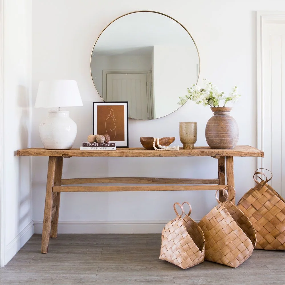 A rough-edged wooden console sits below a round mirror beside pale accessories and woven baskets